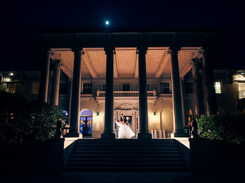 A bride and groom standing in front of a mansion at night.