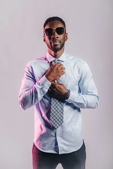 DJ Rakim in sunglasses and a tie posing on a gray background.